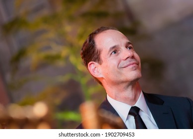An adult white male at a dinner gala smiling with joy as he looks on. He is wearing a dark suit and tie. Foregroune and background.