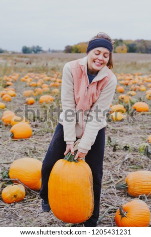 Adult woman (30s) attempts to pick up a giant pumpkin from a pumpkin patch. Smiling and laughing and having fun