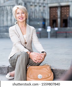 Adult Woman 50-60 Years Old Is Sitting In Time Walking In Old City.