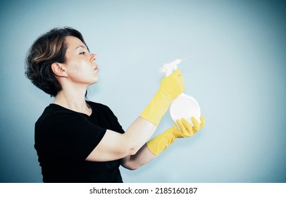 An Adult Woman In A Black T-shirt Splashes With A White Spray Bottle On The Background. Vintage Portrait. Portrait Of Woman Cleaning. Woman In Yellow Cleaning Gloves