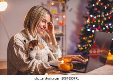 Adult woman in cozy white knitted sweater with dog using laptop  in living room with Christmas tree. Portrait of carefree happy middle aged female in house in christmas - Powered by Shutterstock