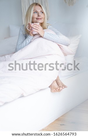 Adult woman with a cup of tea sitting barefooted in bed and looking away with a smile
