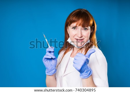 adult woman doctor in protective mask and white medical dressing gown holds syringe and lifts thumb