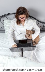 Adult Woman With A Dog With Glasses, Working At Home Working At Home In Her Pijama With A Laptop And A Mobile Phone. Work From Home Concept