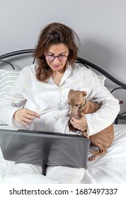 Adult Woman With A Dog With Glasses, Working At Home Working At Home In Her Pijama With A Laptop And A Mobile Phone. Work From Home Concept