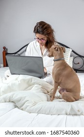 Adult Woman With A Dog With Glasses, Working At Home Working At Home In Her Pijama With A Laptop And A Mobile Phone. Work From Home Concept