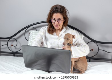 Adult Woman With A Dog With Glasses, Working At Home Working At Home In Her Pijama With A Laptop And A Mobile Phone. Work From Home Concept