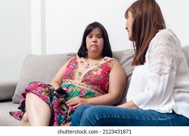 Adult Woman With Down Syndrome Sitting Looking At The Camera Talking With Her Mom At Home. Domestic Life Of A Person With A Disability Without Interaction And Communication Problems.