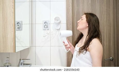 Adult woman dries wet hair with hairdryer and merrily looks in mirror. Lady dances and sings during morning routine after taking shower