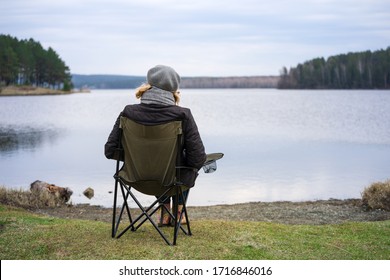 An Adult Woman Enjoys Nature, Sitting In A Camp Chair On The Bank Of A Forest Pond In Early Spring. The Concept Of A Healthy Lifestyle For Older People.