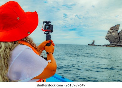 Mujer adulta filmando en barco con un GoPro, de vuelta a la cámara, formaciones rocosas costeras de reserva protegida en la isla de Coronado en el fondo, Parque Nacional Marino de la Bahía de Loreto, Baja California Sur, México