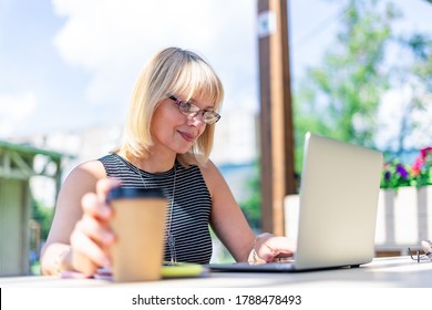 Adult Woman In Glasses Having Video Call With Laptop Outside In Park. Happy And Smiling Senior Working And Drinking Coffee. Using Computer. Distance Learning Online Education And Online Shops.