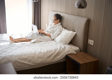 Adult woman in glasses wearing white bathrobe working with laptop in hotel room - Powered by Shutterstock