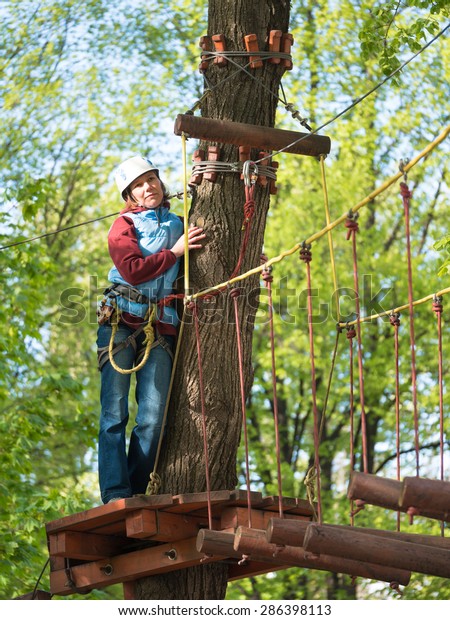 Adult woman in a helmet and with a safety\
system standing beside a tree and the suspended rope bridge on a\
blurred background