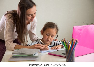 Adult woman helping ethnic girl to do homework assignment in notebook near laptop during studies at home