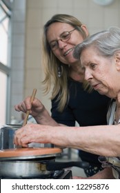 Adult Woman And Her Elderly Mother Cooking Together In Their Kitchen. The Older Lady Is Lifting Up The Lid Of A Pot And Looking Inside.