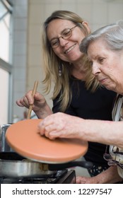 Adult Woman And Her Elderly Mother Cooking Together In Their Kitchen. The Older Lady Is Lifting Up The Lid Of A Pot And Looking Inside.