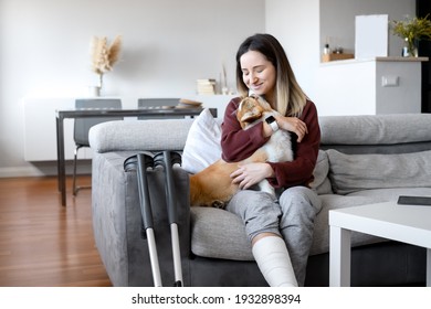 Adult Woman In Her Late Twenties On Couch At Home With Crutches And Orthopedic Plaster Caress The Dog. Fracture Of The Leg Or Foot. Concept Of Rehabilitation And Healing. Orthopedics And Traumatology.