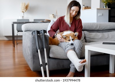 Adult Woman In Her Late Twenties On Couch At Home With Crutches And Orthopedic Plaster Caress The Dog. Fracture Of The Leg Or Foot. Concept Of Rehabilitation And Healing. Orthopedics And Traumatology.