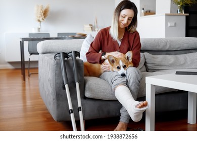 Adult Woman In Her Late Twenties On Couch At Home With Crutches And Orthopedic Plaster Caress The Dog. Fracture Of The Leg Or Foot. Concept Of Rehabilitation And Healing. Orthopedics And Traumatology.