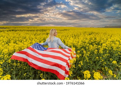 Adult Woman Holding American Flag With Pole, Stars And Stripe In A Yellow Rapeseed Field. USA Flag Fluttering In The Wind