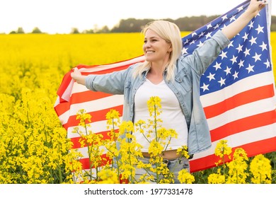 Adult Woman Holding American Flag With Pole, Stars And Stripe In A Yellow Rapeseed Field. USA Flag Fluttering In The Wind