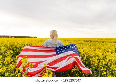 Adult Woman Holding American Flag With Pole, Stars And Stripe In A Yellow Rapeseed Field. USA Flag Fluttering In The Wind