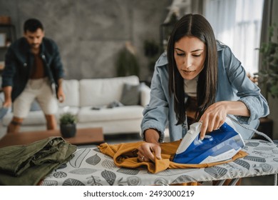 Adult woman iron the wardrobe on the iron board while man clean dust - Powered by Shutterstock