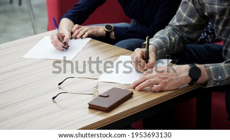 An adult woman and a man, sitting at a table in the office, fill out documents or forms. The concept of a marriage contract, agreement or divorce proceedings. Without a face.