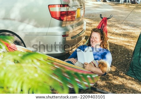 Adult woman relaxing and reading book while lying in hammock with cockapoo puppy near motorhome on camping trip. Female living on camper car with animals and travel the world. Caravan car Vacation.