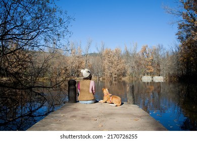 Adult Woman Sitting Next To Pet Dog On Lake Dock In Forest