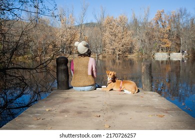 Adult Woman Sitting Next To Pet Dog On Lake Dock In Forest