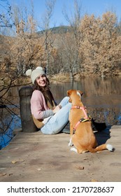 Adult Woman Sitting Next To Pet Dog On Lake Dock In Forest