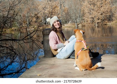 Adult Woman Sitting Next To Pet Dog On Lake Dock In Forest