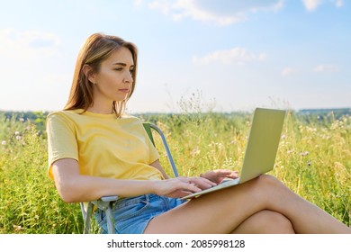 Adult Woman Sitting On An Folding Camping Chair Using A Laptop, In A Meadow With Wild Herbs