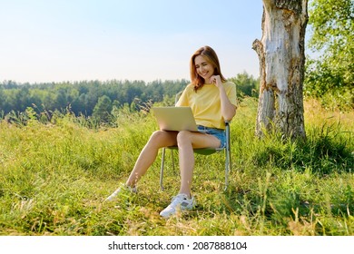 Adult Woman Sitting On An Folding Camping Chair Using A Laptop, In A Meadow With Wild Herbs