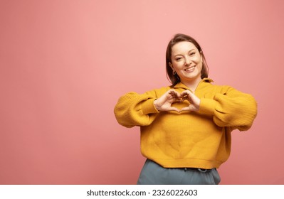 Adult woman smiling, showing hand heart gesture over chest and staring at camera, making love confession, standing against pink background. - Shutterstock ID 2326022603
