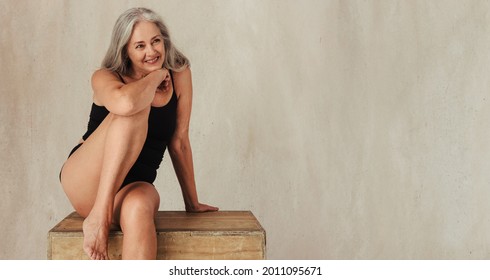 Adult woman smiling while posing in her natural body. Confident woman in black underwear embracing her aging body. Mature woman sitting alone against a studio background.