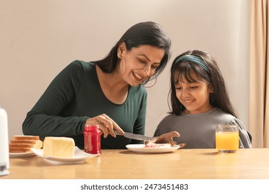 AN ADULT WOMAN SPREADING JAM ON BREAD FOR HER DAUGHTER SITTING BESIDE - Powered by Shutterstock