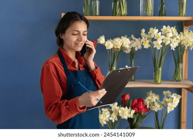 adult woman talks on phone while holding a clipboard working in her flower shop with various flowers. Copy space - Powered by Shutterstock