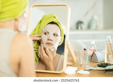 Adult woman in towel turban on head applying hydrating face mask during daily self-care routine after shower at home.. - Powered by Shutterstock