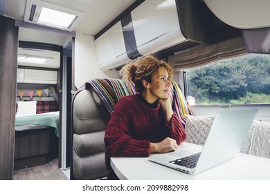 Adult woman use laptop computer inside a camper van recreational vehicle sitting at the table with bedroom in background and nature park outside the windows. Concept of travel and remote worker people