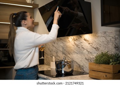 An adult woman using the control panel of the modern cooker hood. Kitchen room with cozy interiors and contemporary built-in appliances.