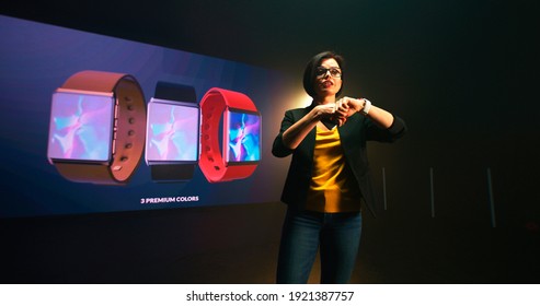 Adult Woman Walking On Stage And Starting To Explain Features Of New Smart Watch Connectable With Smartphone On Stage Against LED Screen