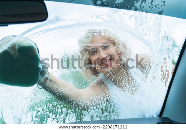 An adult woman washes the windshield of a\
car, a view from inside the car\
interior