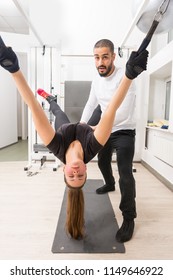 Adult Woman Wearing Black Tee Shirt Held Above Yoga Mat By Black Leather Straps With Instructor Standing Beside Her