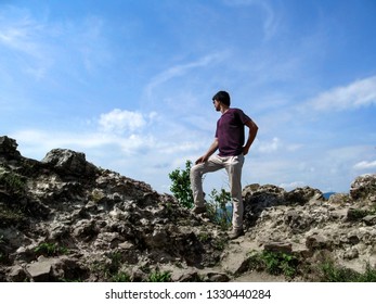 The adult young man climbed a rock and looks into the distance in a winner pose. Dark-haired slim guy in a T-shirt, white pants and shoes stands on top of the mountain - view from the back