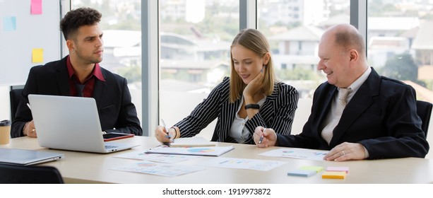 Adults Caucasian Business 2 Men And 1 Woman Are Meeting To Discuss The Marketing Plan And Have Notebook, Laptop, Tablet Put On Table And Whiteboard Are Backward Of Them With Sunlight In The Morning.