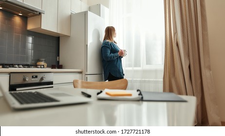 Advantages Of Working From Home. Young Woman In Casual Clothes Drinking Coffee Or Tea, Looking In The Window While Standing In The Kitchen At Home. Taking A Break. Freelance. Home Office