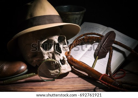 Adventure and archeology concept. Skull with fedora hat, bullwhip, book, quill, shoe, mortyr and magnifying glass on wooden table and black background. Still life